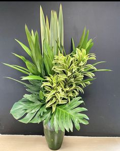 a green vase filled with lots of plants on top of a wooden table next to a gray wall