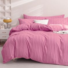 a bed with pink comforter and pillows in a white room next to a lamp