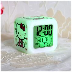 an alarm clock with hello kitty on it is sitting on a white tablecloth next to a pink flower