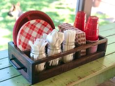 a wooden tray with red and white plates, napkins and cups in it on a picnic table