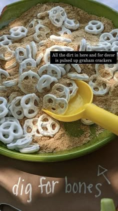 a green bowl filled with lots of white letters on top of sand and a yellow spoon