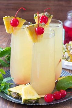 two glasses filled with pineapple and cherries on top of a plate next to fruit