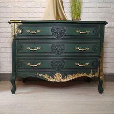 a green and gold painted dresser in front of a white brick wall with a potted plant on top