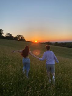 two people holding hands walking through tall grass at sunset with the sun setting behind them