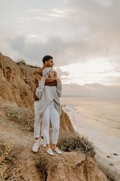 a man and woman standing on top of a cliff next to the ocean with their arms around each other