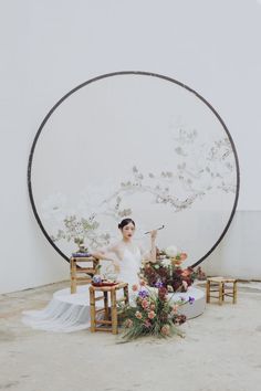 a woman sitting in front of a round mirror with flowers on the table next to her