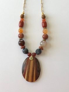 Lightweight oval wooden pendant has different shades of brown. Necklace has different shapes and sizes of stone beads in earthtone colors and hangs from a gold plated chain with oblong links and has a gold plated lobster clasp. Earth Tone Jewelry, Different Shades Of Brown, Peace Necklace, Wooden Bead Necklaces, Brown Necklace, Wooden Pendant, Shades Of Brown, Jasper Pendant, Leaf Charms