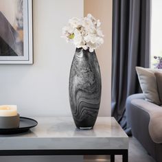 a black vase with white flowers on a table in front of a couch and window