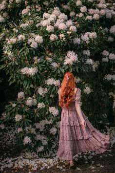 a woman with long red hair is standing in front of a bush full of white flowers