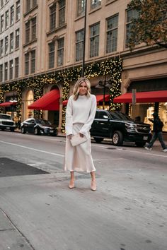 a woman is standing on the street wearing a white sweater and midiskile skirt