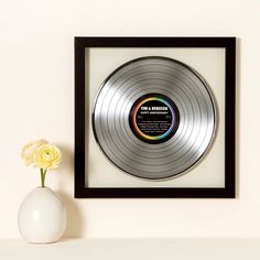 a silver record in a black frame next to a white vase with a yellow flower