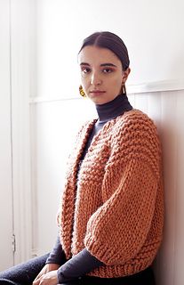 a woman wearing an orange sweater and black pants sitting on the floor in front of a white wall