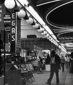 black and white photograph of people walking in an outdoor shopping area with lights hanging from the ceiling