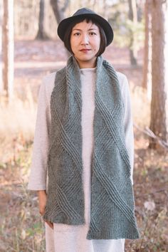 a woman standing in the woods wearing a green knitted vest and black hat with her hands on her hips