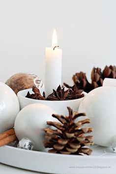 a lit candle surrounded by white ornaments and pine cones on a plate with cinnamon sticks