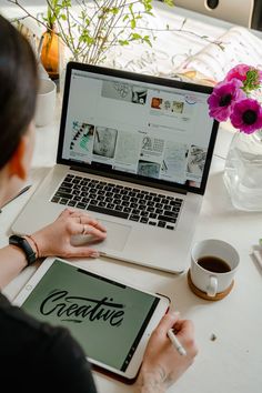 a woman sitting at a table with a laptop and tablet in front of her, writing on the screen