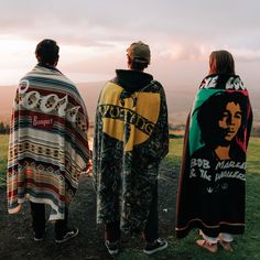 three people standing on top of a grass covered field next to each other wearing blankets