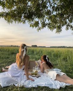 two women in white dresses sitting on a blanket under a tree drinking wine and having a picnic