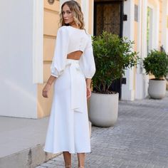 a woman standing on the sidewalk wearing a white dress with cut out shoulders and long sleeves