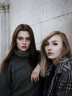 two beautiful young women standing next to each other near a stone wall and looking at the camera