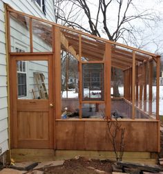 a small wooden greenhouse in front of a house with snow on the ground and bare trees