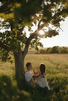 a man and woman sitting under a tree on the grass in front of a field