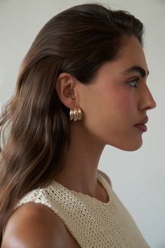 a woman with long brown hair wearing gold earrings and a white top is looking off to the side