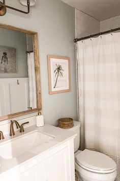 Beachy bathroom decor shows a neutral bathroom with a photo of a palm tree in a sandy-colored frame. Wooden accents, a neutral paint color, and a beige striped shower curtain are perfect for a modern beachy bathroom. White Beach Bathroom Ideas, Simple Beach Bathroom Ideas, Small Coastal Apartment Decor, Blue Beach Bathroom Ideas, Coastal Guest Bathroom Decor, Beach Guest Bathroom Ideas, Beachy Guest Room, Beachy Chic Decor, Boho Beach House Bathroom