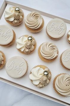 twelve cupcakes with white frosting in a box on a counter top, decorated with the word baby