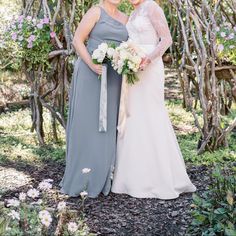 two women standing next to each other in front of some trees and bushes with flowers