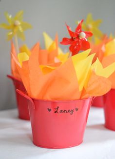 three red buckets filled with paper flowers on top of a white cloth covered table