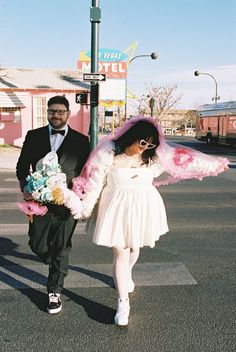 a man and woman are walking down the street with pink feathers on their arms, dressed in white