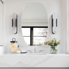 a white bathroom with a large round mirror above the sink and two vases on the counter