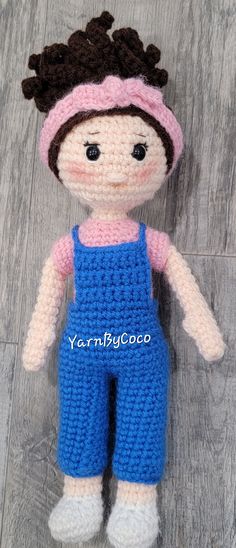 a crocheted doll wearing blue overalls and a pink headband on a wooden floor