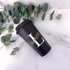 a black coffee cup sitting on top of a white table next to green leaves and ivy