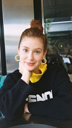 a woman sitting at a table with her hand on her chin and looking into the camera