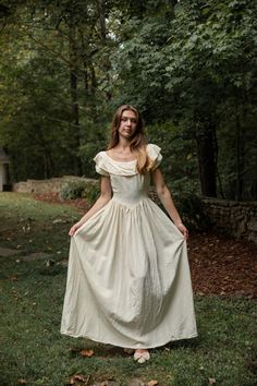 a woman in a white dress standing on grass with trees and bushes behind her, looking at the camera