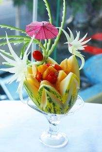 a glass bowl filled with cut up fruit and flowers on top of a table next to a window