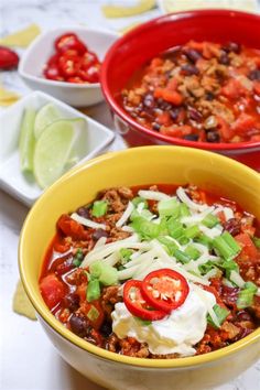 two bowls of chili with sour cream, tomatoes and green onions next to limes