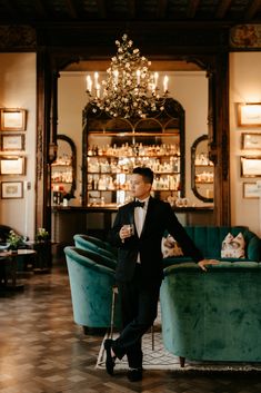 a man in a tuxedo standing next to a green couch and chandelier