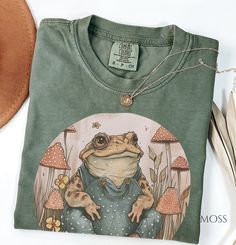 Title: "Retro Cottagecore Toad Comfort Color Shirt | Vintage-inspired Nature Tee| Botanical Shirt Description: Step into a whimsical world of nostalgia with our Retro Cottagecore Toad Comfort Color Shirt. Inspired by the enchanting allure of cottagecore aesthetics and vintage charm, this tee features a delightful depiction of a toad in a retro style, capturing the essence of nature's beauty and simplicity. Crafted on Comfort Color 1717, our shirt offers unparalleled comfort and quality, ensuring Green Goblincore Cotton Top, Green Cotton Goblincore Top, Retro Cottagecore, Nature Tees, Etsy Ideas, Botanical Shirt, Frog And Toad, Color Shirt, Comfort Color