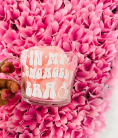 someone is holding up a glass cup with pink flowers in the background and writing on it