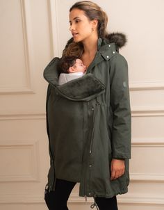 a woman is holding a baby in a green coat and black leggings while standing against a white wall