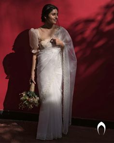 a woman wearing a white sari and holding a bouquet in front of a red wall