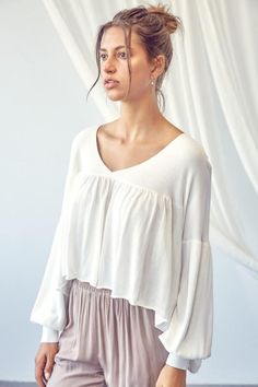 A slightly cropped knit long sleeve white top featuring a v neckline, dropped shoulders, balloon sleeves that cuff at ends, and a high low hem. Also available in mocha. Model Measurements: Height 5'9", Bust 32, Waist 24, Hips 34. Wearing a size Small.