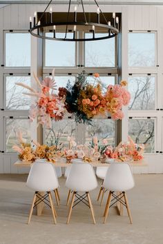 a dining table with white chairs and flowers on it in front of a large window