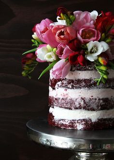 a red velvet cake with white frosting and pink flowers on top, sitting on a silver pedestal