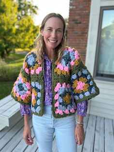 a woman standing on a porch wearing a colorful crochet jacket and smiling at the camera