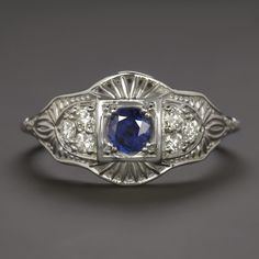 This beautiful and unique ring features a richl blue sapphire set in a diamond studded vintage style design that will never go out of style! The sapphire has a richly saturated blue hue that contrasts strikingly with the 14k white gold setting. Utterly classic and romantic, the antique style setting is richly textured with intricate filigree and milgrain. The effect is chic but also dreamy and softly feminine, a perfect choice for a timelessly stylish right hand ring or alternative engagement ri Blue Platinum Art Deco Diamond Ring, Classic Blue Diamond Ring, Art Deco Blue Diamond Ring, Blue Platinum Jewelry With Single Cut Diamonds, Platinum Jewelry With Blue Single Cut Diamonds, Blue Diamond Art Deco Ring, Blue Single Cut Diamond Jewelry In Platinum, Blue Art Deco Diamond Ring, Art Deco Blue Diamond Ring With Center Stone