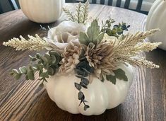 a white pumpkin decorated with flowers and greenery sits on a wooden table in front of two white pumpkins